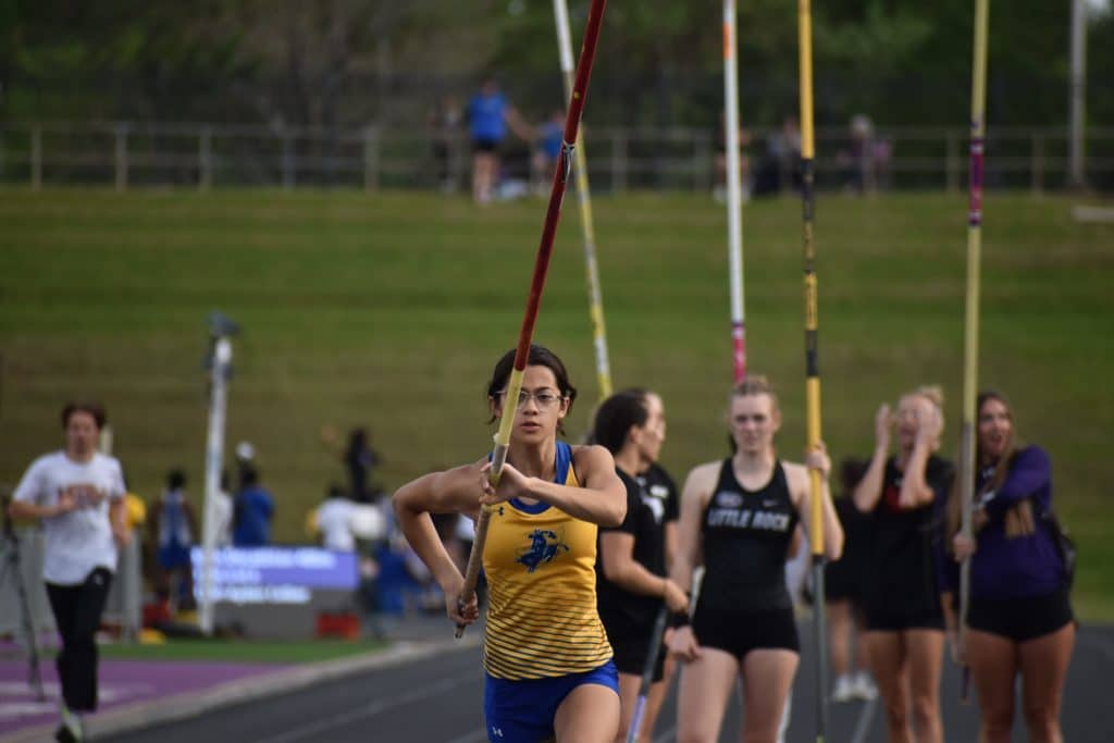 A Mulerider pole vaulter plants her pole as teammates and competitors look on